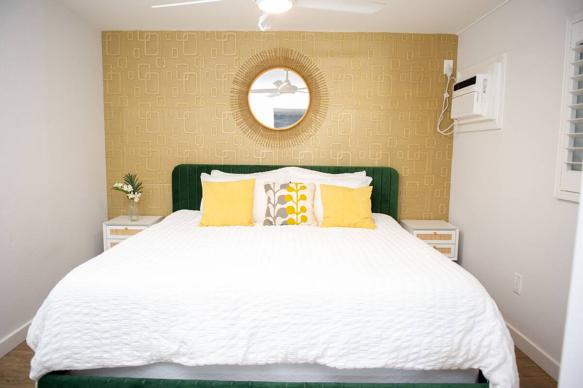 A neatly made bed with white bedding and yellow pillows, a green headboard, and a wall-mounted round mirror.
