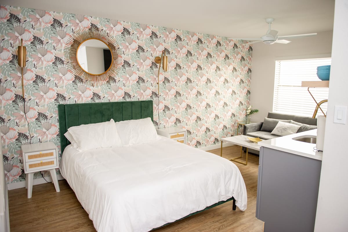 A bedroom with a green headboard, a white bed, a round mirror, and a floral wallpaper pattern.