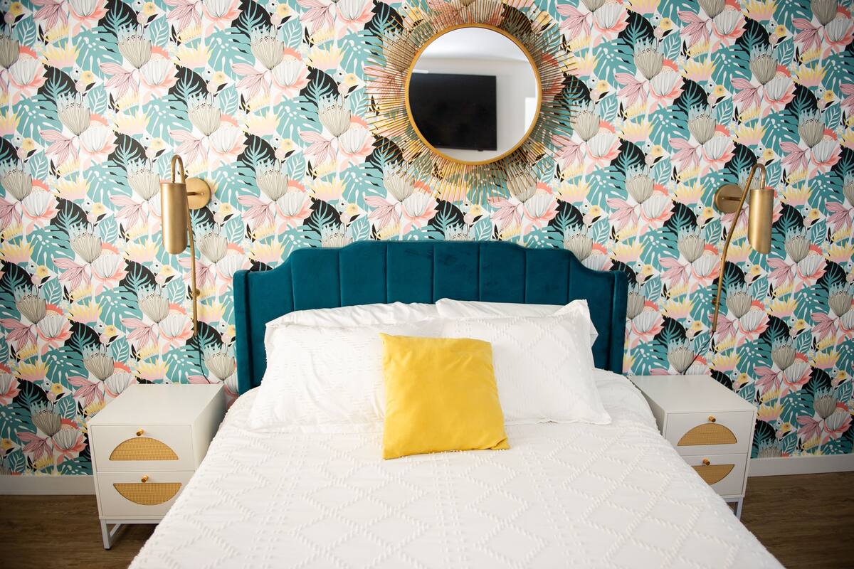 A bedroom with a floral wallpaper, a teal upholstered headboard, a white bed with a yellow pillow, and two bedside tables with gold lamps.