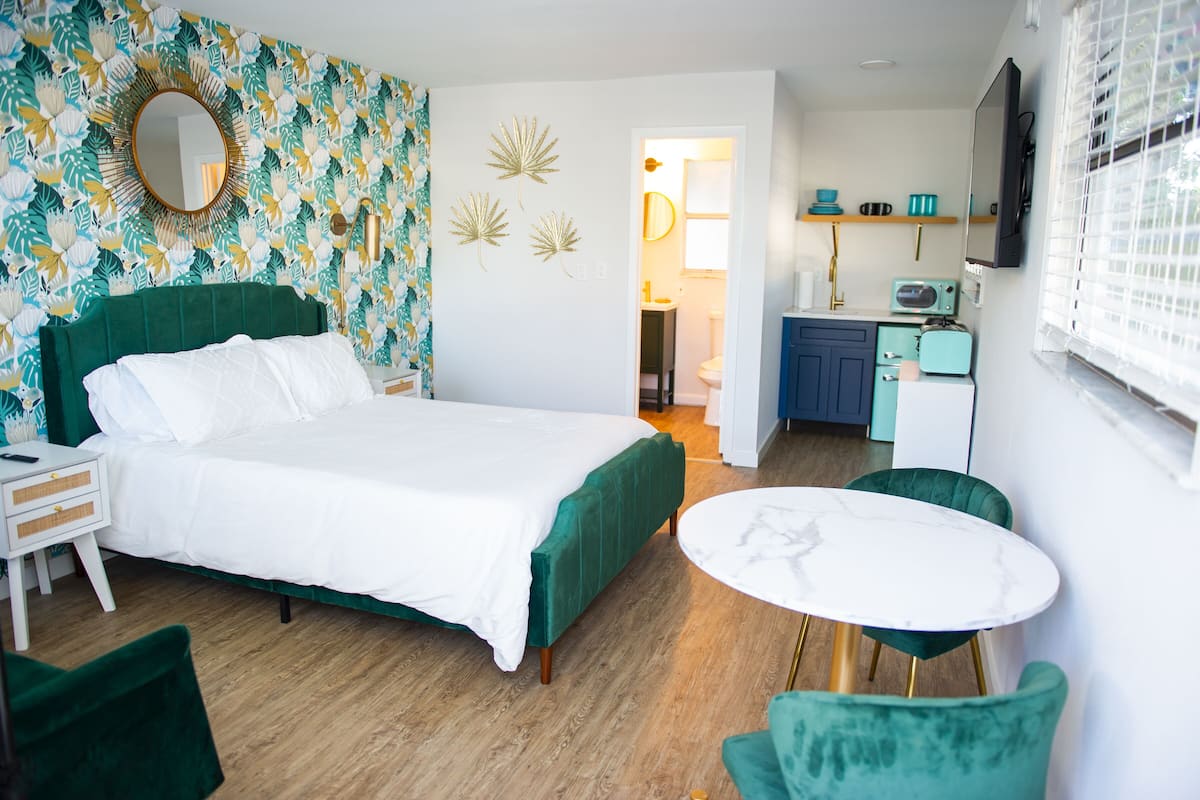 A hotel room with a bed, a desk, a chair, a small table, and a kitchenette area.