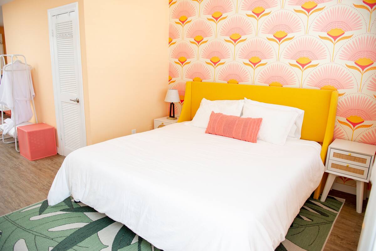 A neatly made bed with white and pink bedding, a yellow headboard, and a patterned wall.