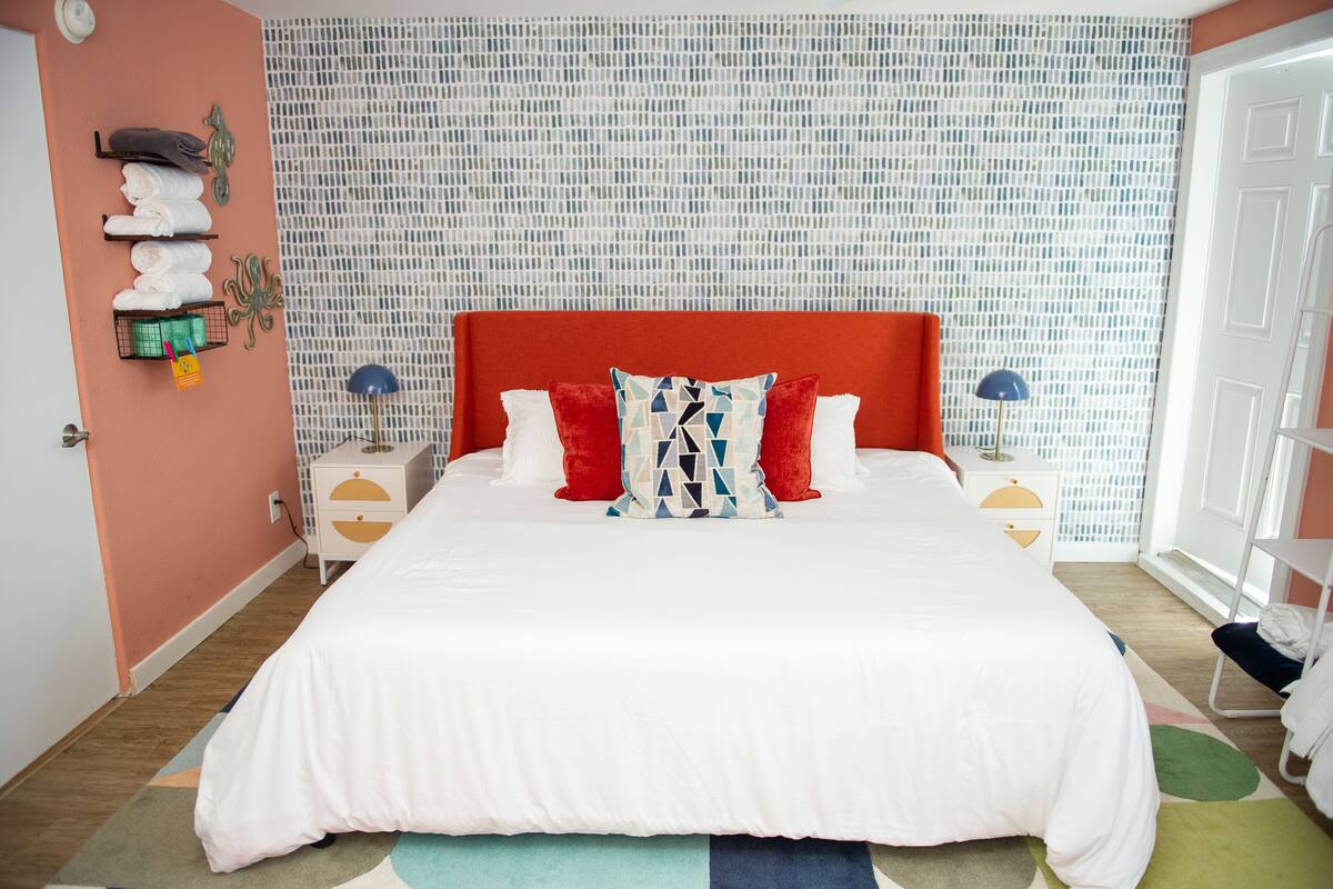 A neatly made bed with a red headboard, white bedding, and two red pillows. There are two bedside tables with lamps, a wall-mounted shelf with towels, and a patterned wall behind the bed.