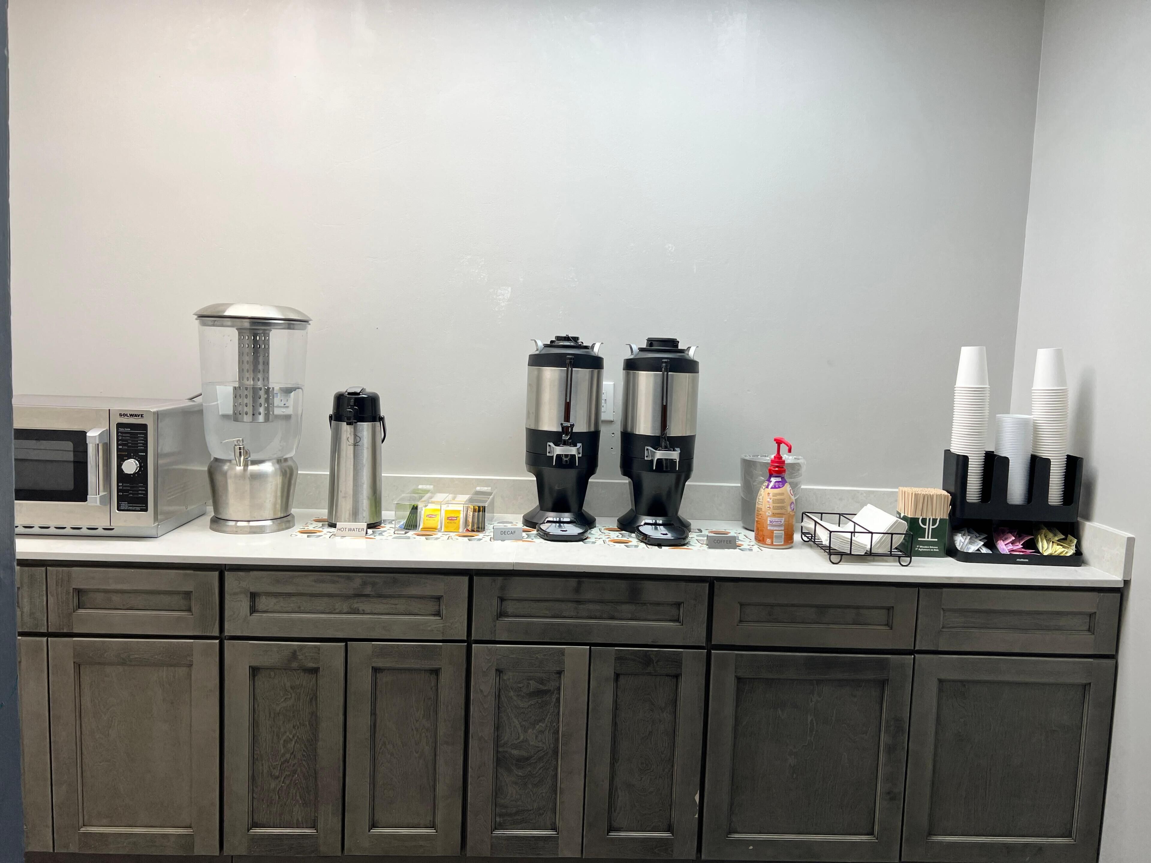 A kitchen with a microwave, hot water dispenser, coffee maker, and coffee station.