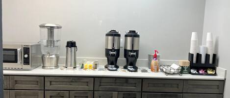 A kitchen with a microwave, hot water dispenser, coffee maker, and coffee station.