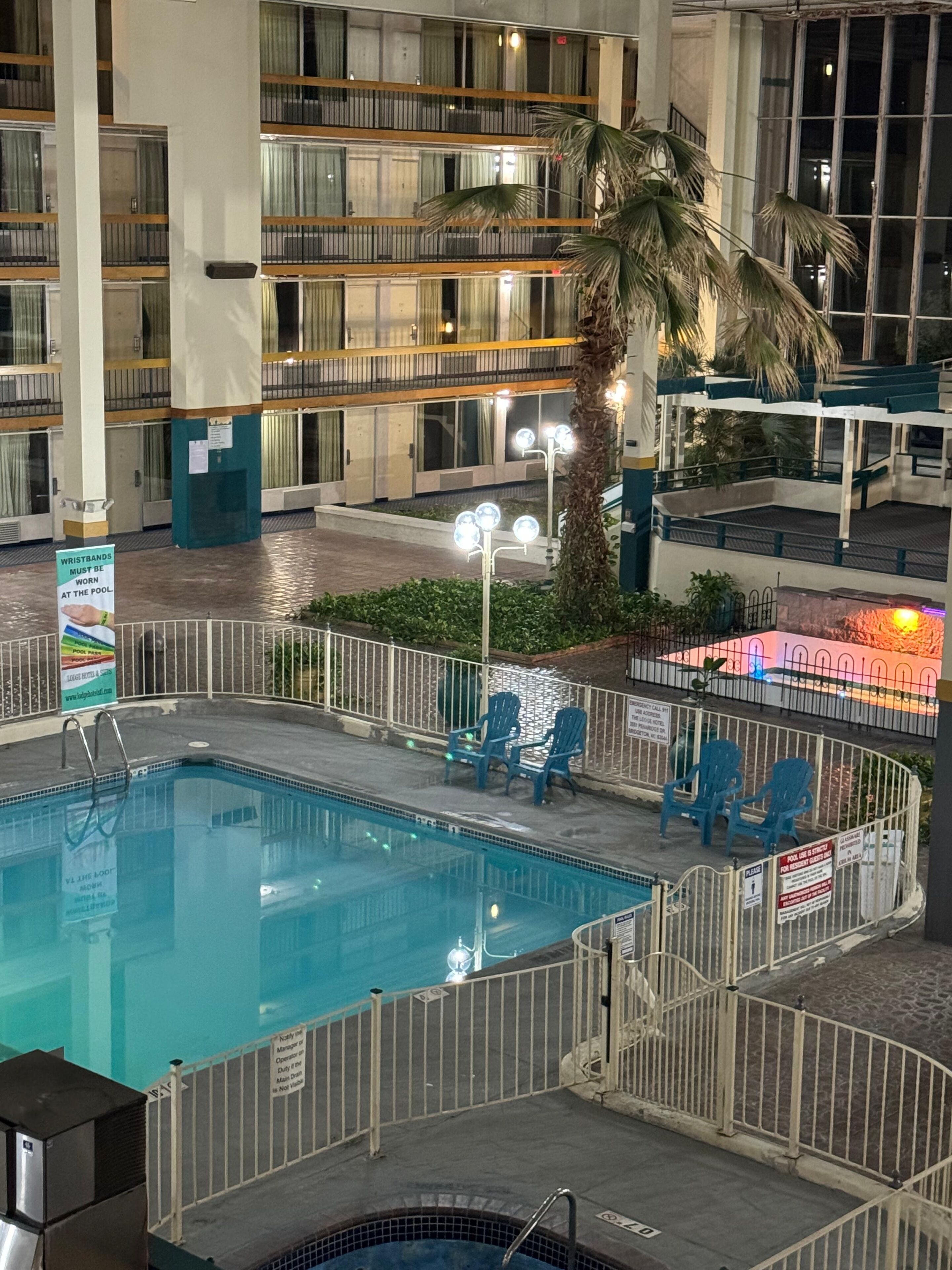 An indoor swimming pool with a tiled deck, surrounded by a white fence. There are blue chairs and a palm tree near the pool.