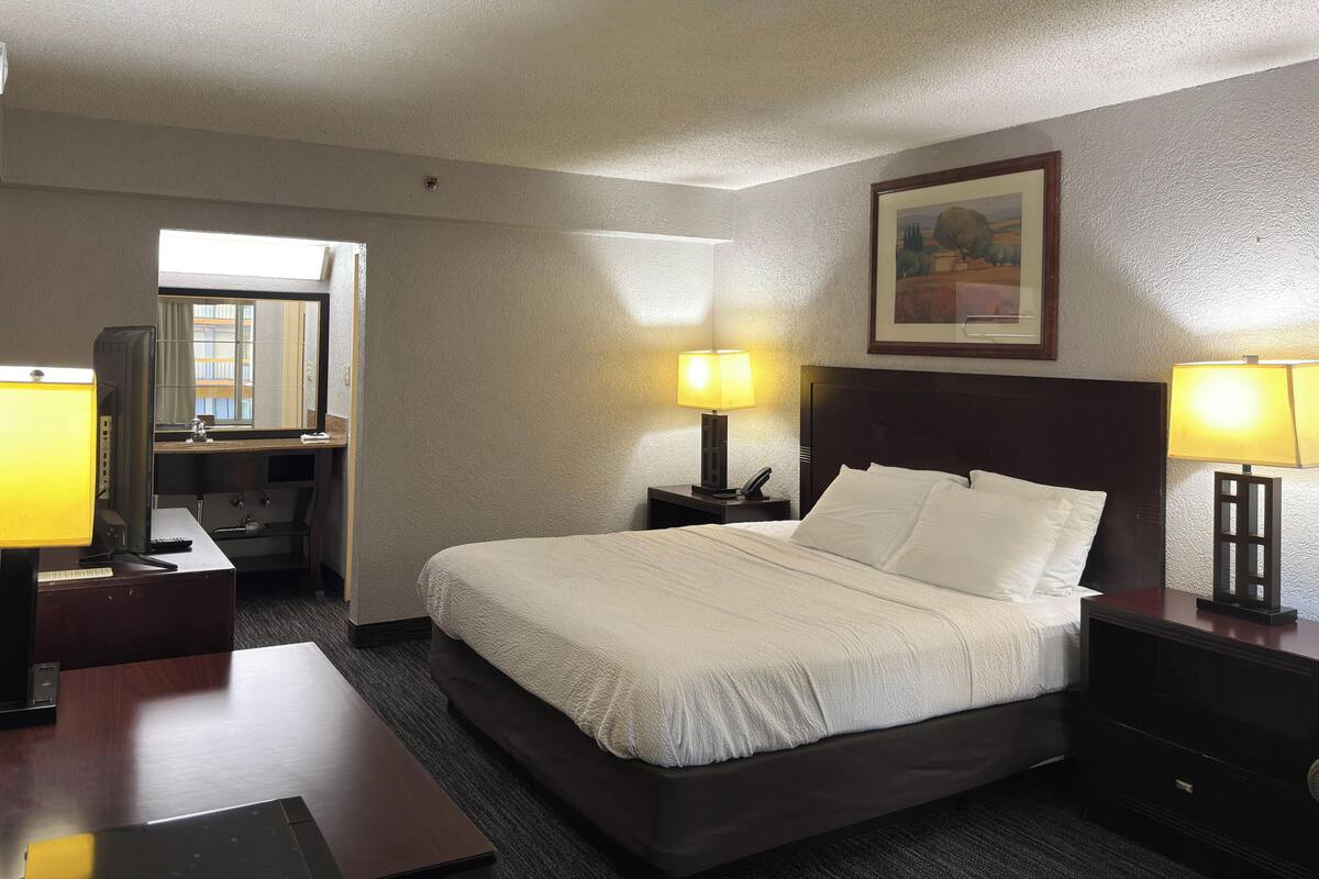 A hotel room with a bed, two bedside tables with lamps, a desk with a computer, a chair, and a window with a view of buildings.