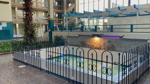 An indoor swimming pool with a stone wall and a water feature, surrounded by a metal fence. - The Lodge Hotel and Banquets (Bridgeton)