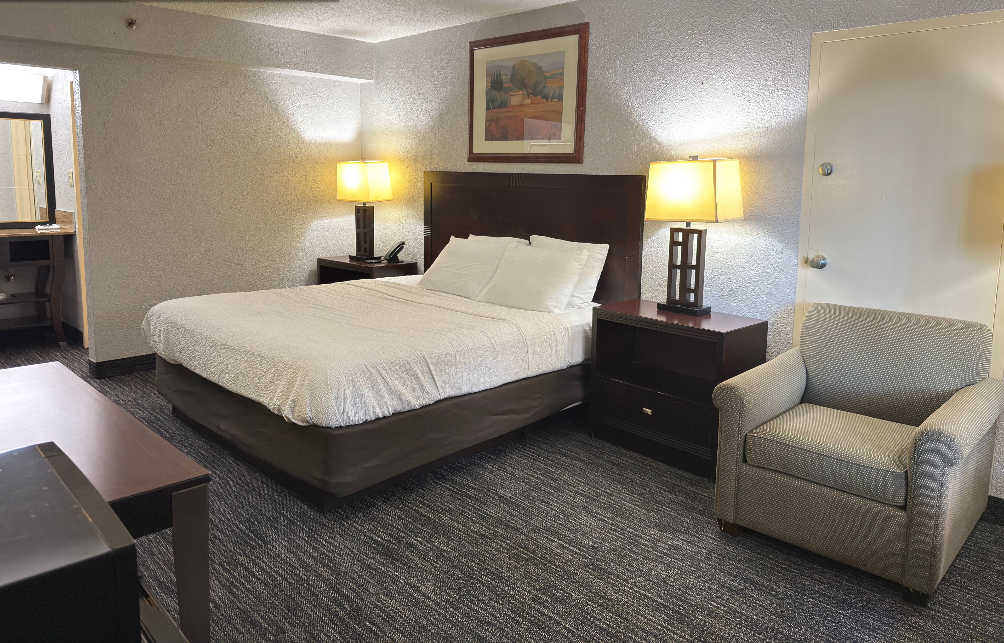A hotel room with a bed, two bedside tables with lamps, a chair, and a door.