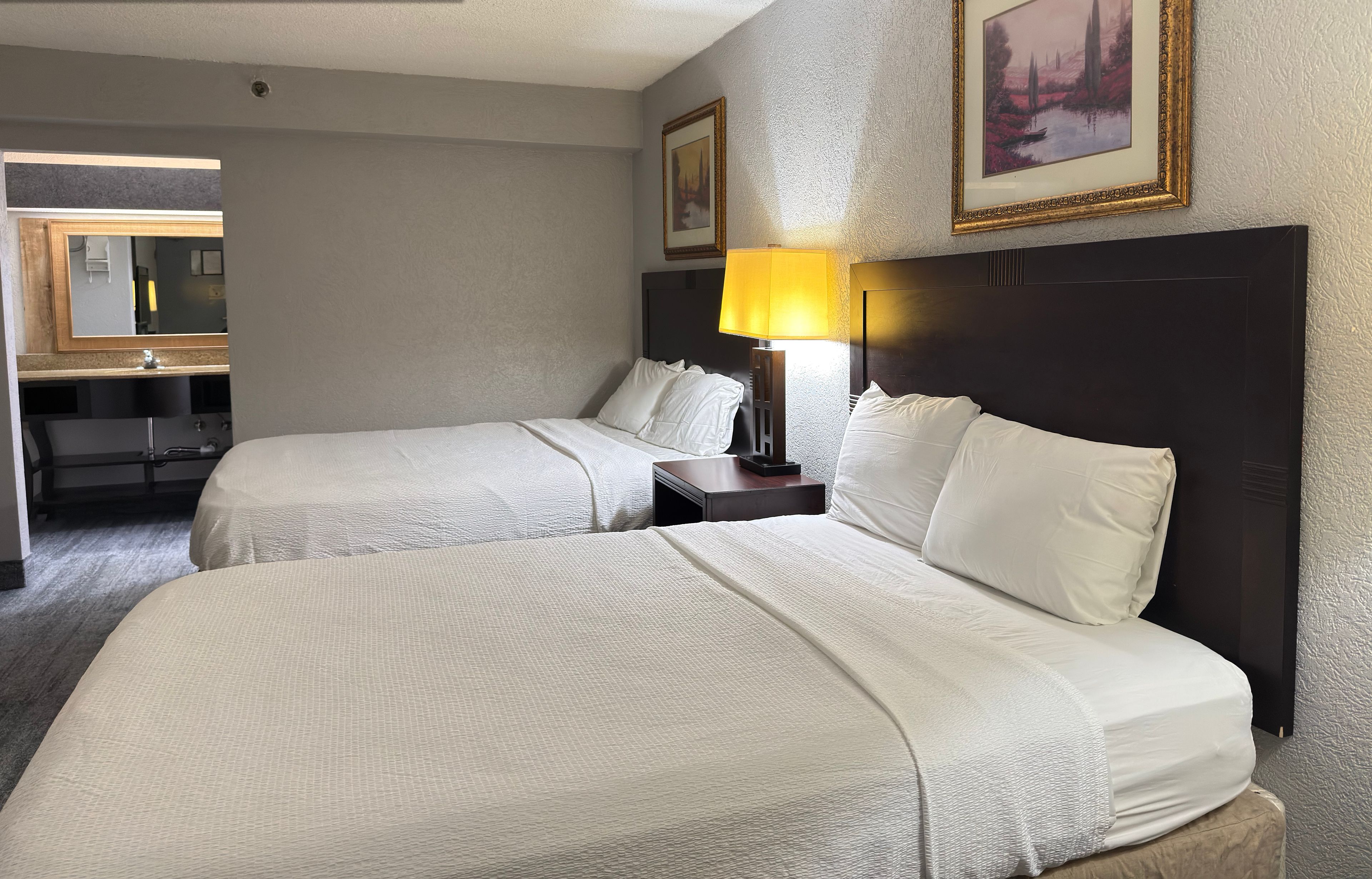 Een hotelkamer in renovatie met twee bedden, een nachtkastje met een lamp, een spiegel en ingekaderde foto's aan de muur.