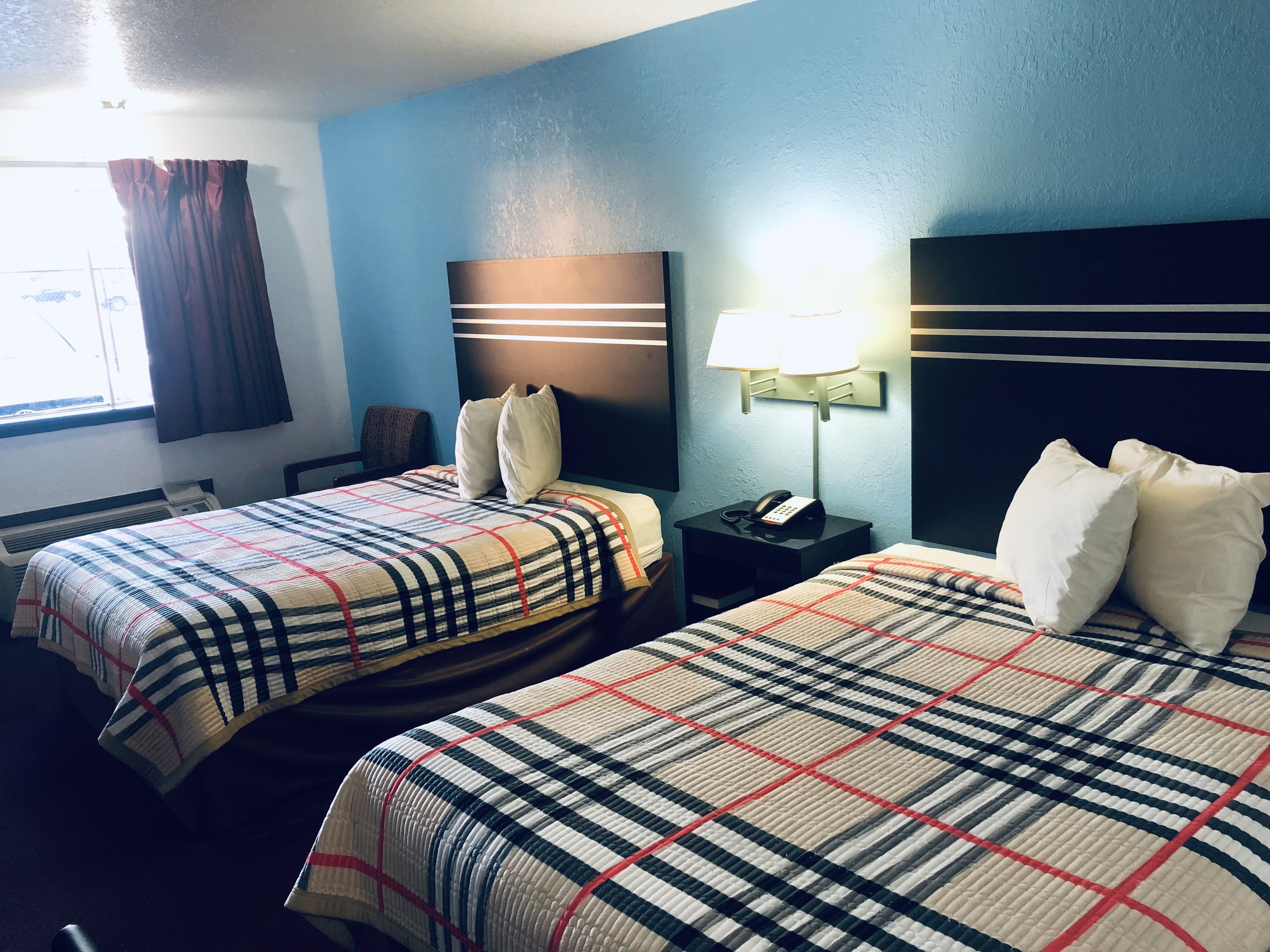A hotel room with two beds, each with a plaid bedspread, a bedside table with a lamp, and a wall-mounted phone.