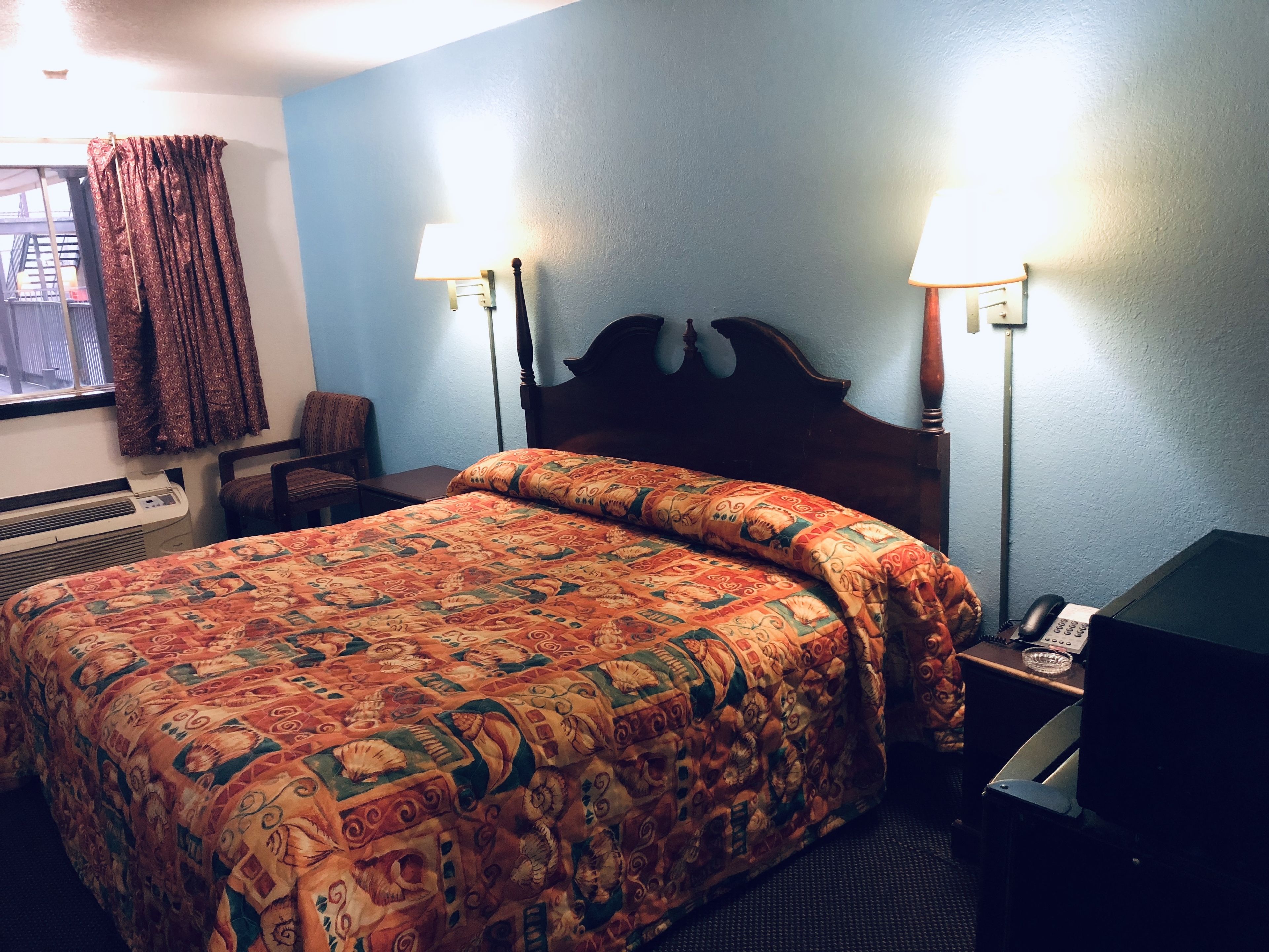 A hotel room with a large bed, two bedside lamps, a chair, a desk with a computer, and a telephone.