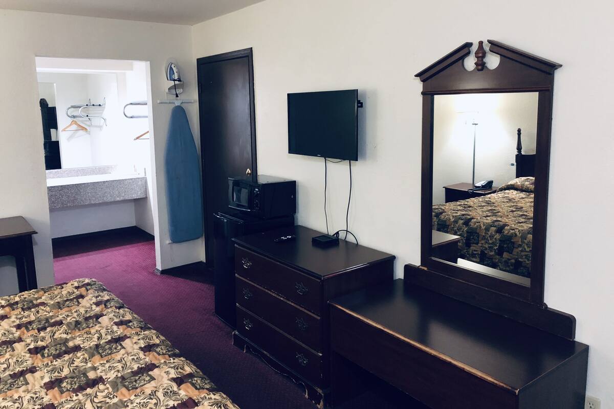 A hotel room with a bed, dresser, mirror, television, microwave, iron, and a desk with a chair.