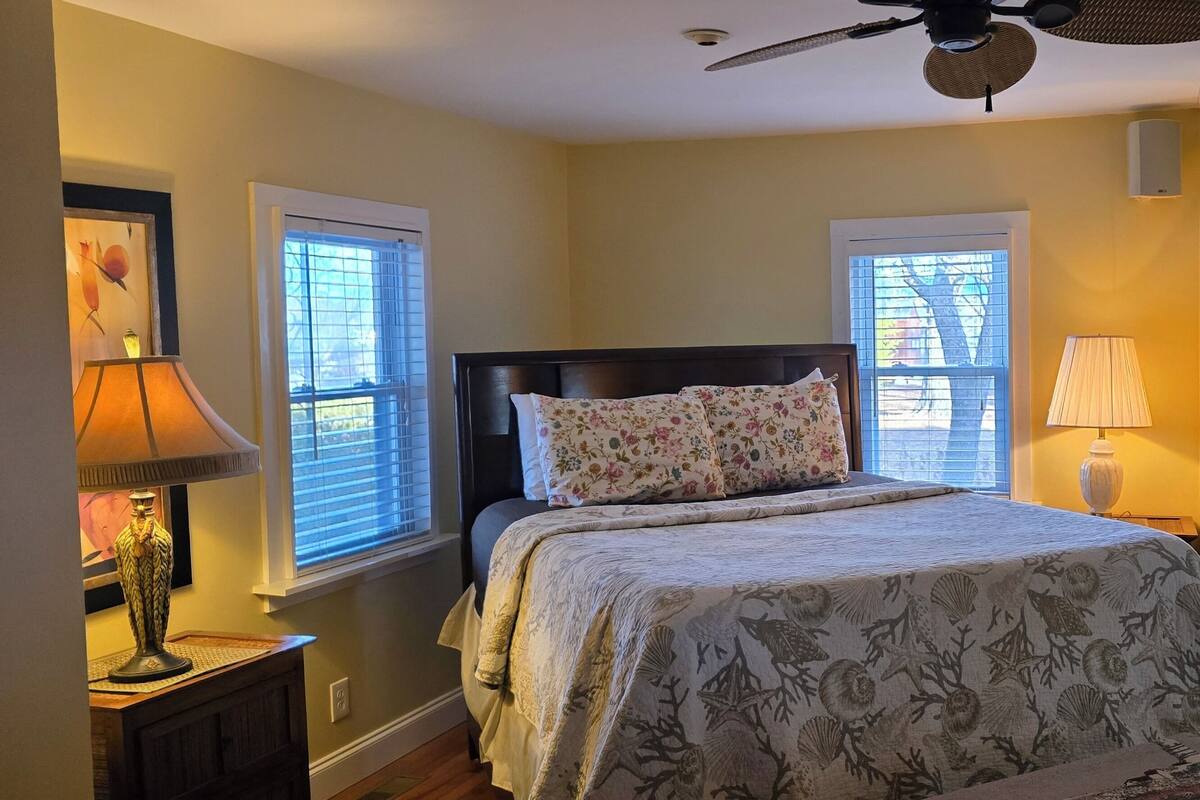 Une chambre à coucher avec un lit, deux lampes de chevet, un ventilateur de plafond et des fenêtres équipées de stores.