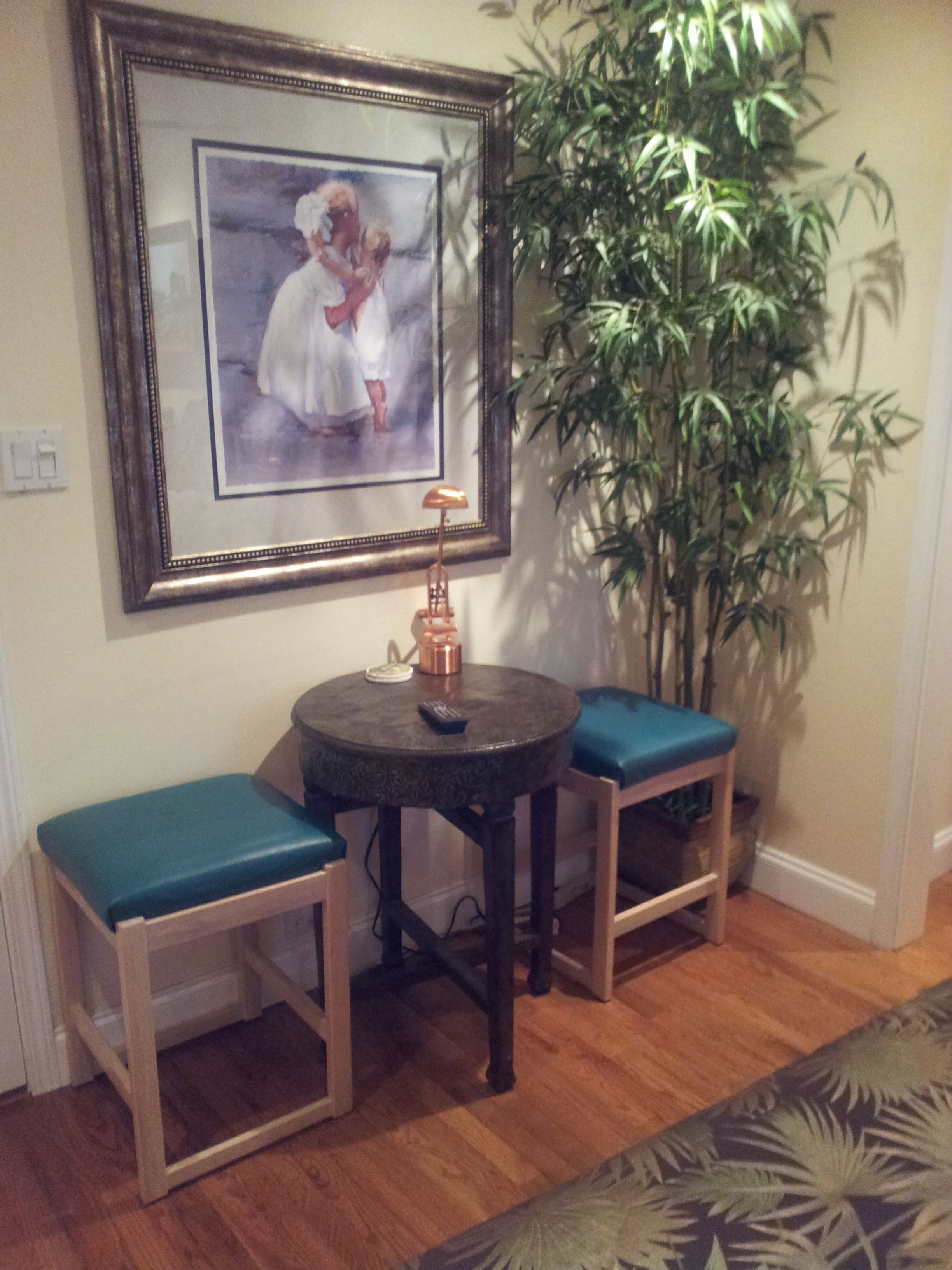A room with a framed painting, a small round table with a lamp, two stools, a large potted plant, and a patterned rug.