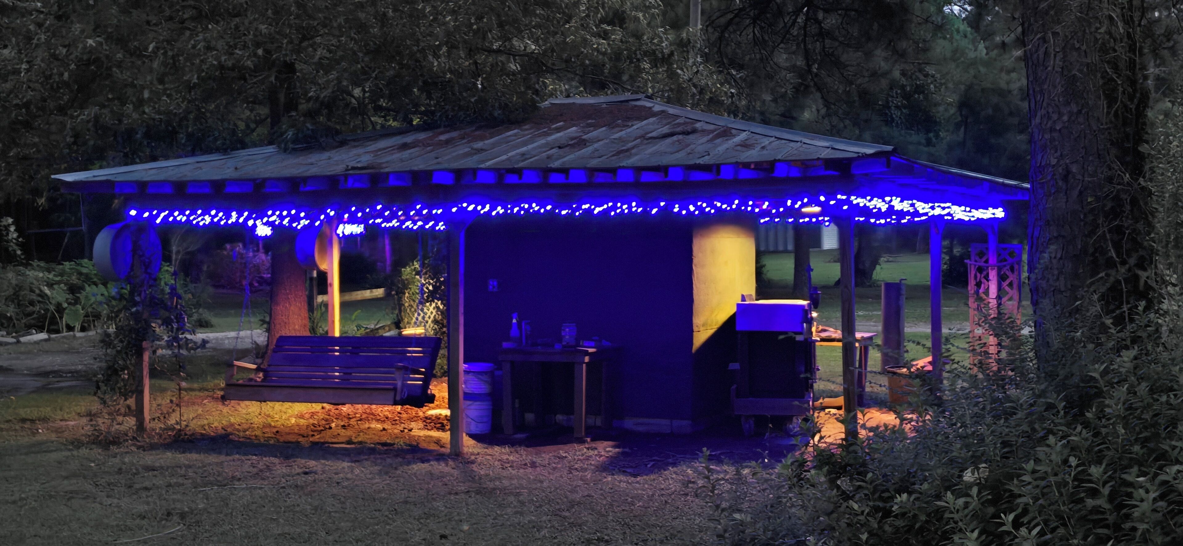 Ein rustikaler Kiosk mit Holzboden und blauen Lichterketten, umgeben von Bäumen.