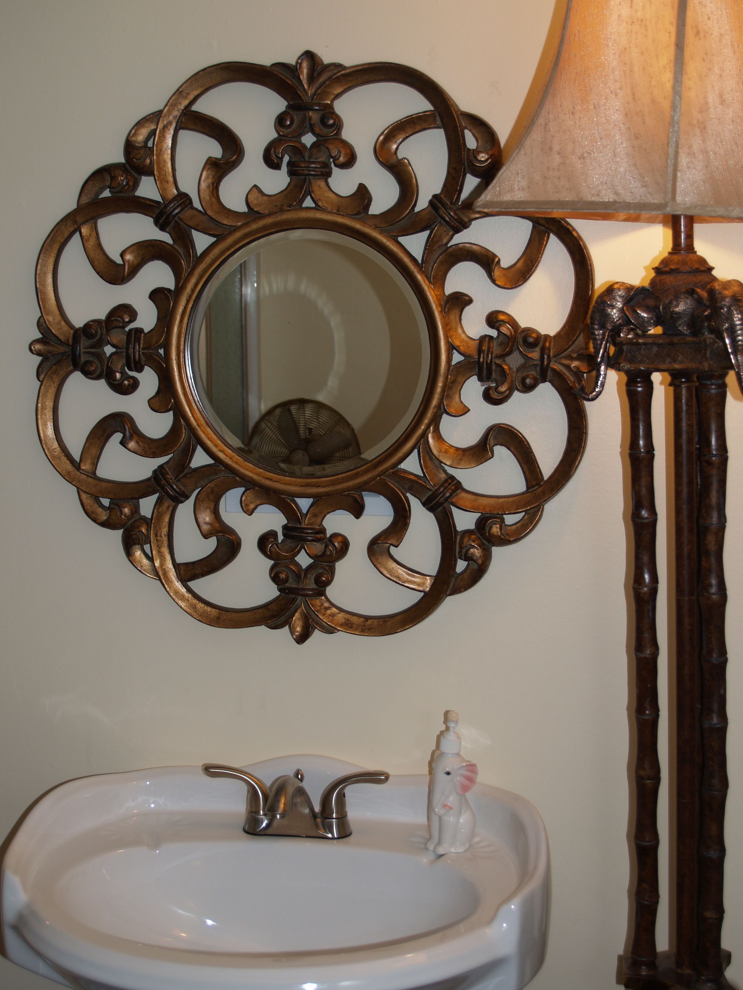 Une salle de bain avec un lavabo blanc, un miroir doté d’un cadre orné et une lampe.
