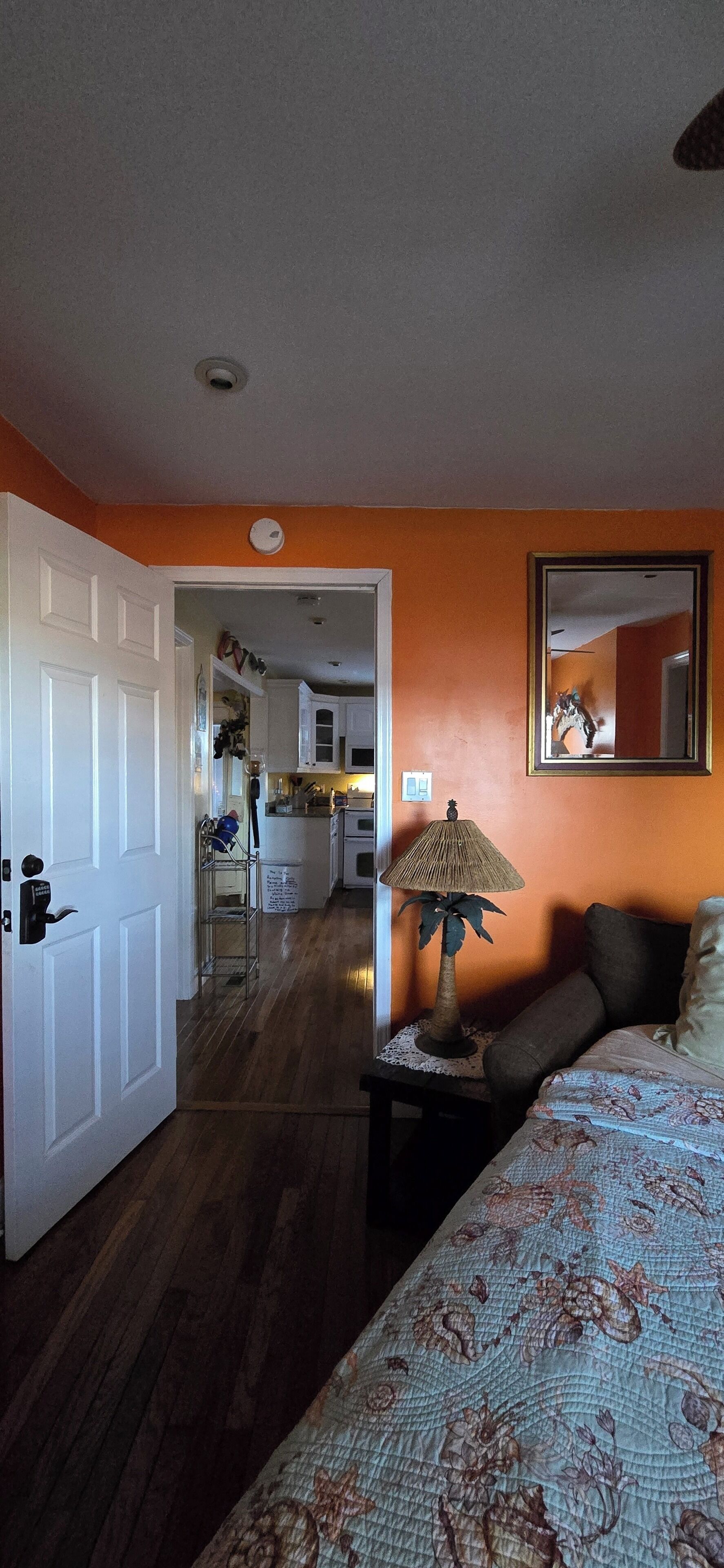 Un dormitorio con cama, una mesita de noche con lámpara, una puerta abierta que da a una cocina y un espejo en la pared.
