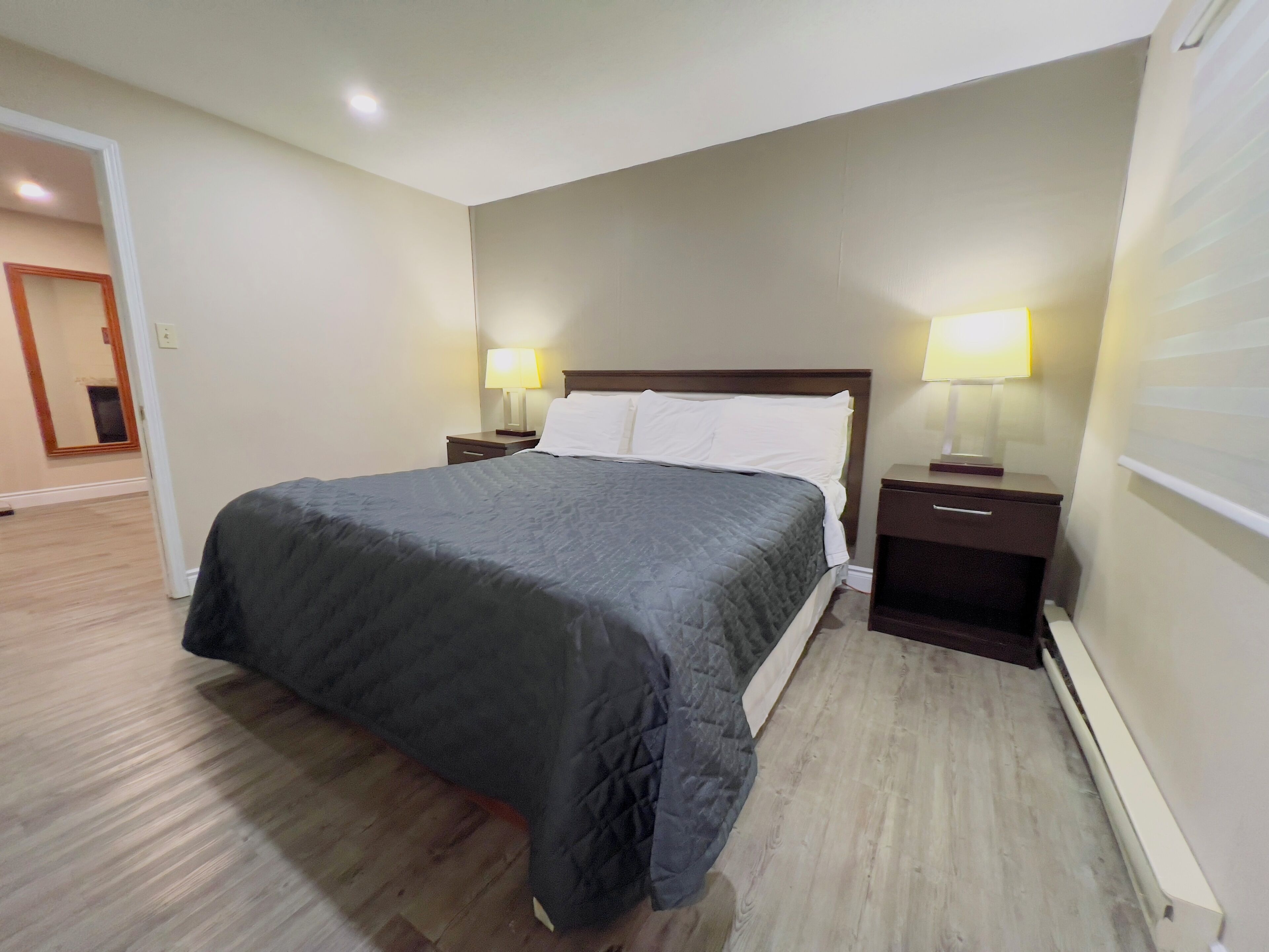 Une chambre d’hôtel avec un lit, deux tables de chevet avec des lampes, un plancher en bois et une porte donnant sur une autre pièce.