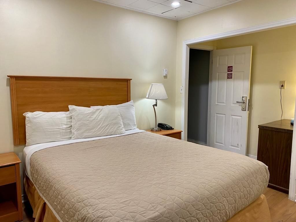 Cama bem arrumada com colcha bege, cabeceira de madeira, luminária de cabeceira e mesa de apoio com um telefone.