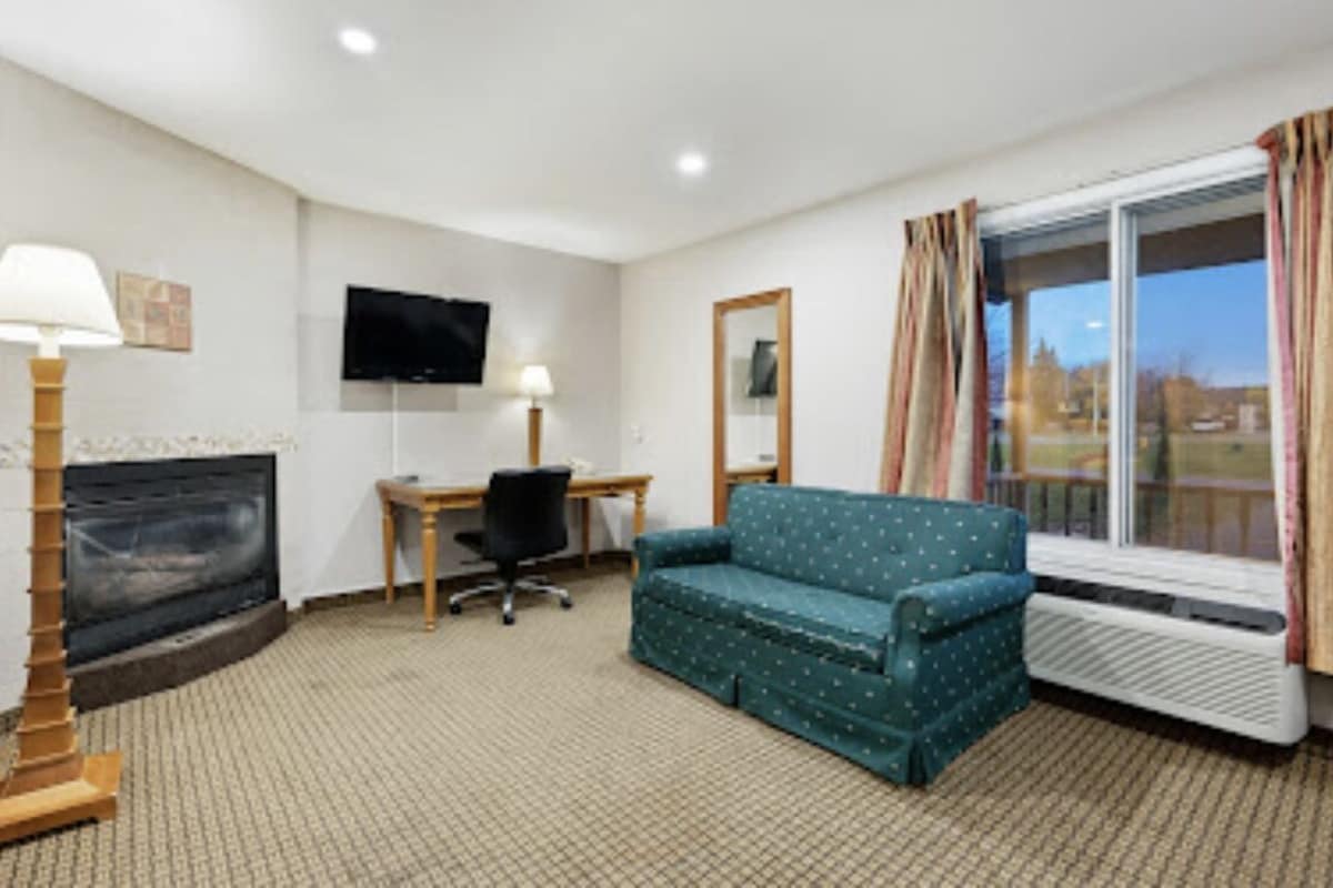 A room with a fireplace, a television mounted on the wall, a desk with a chair, a sofa, and a window with curtains.