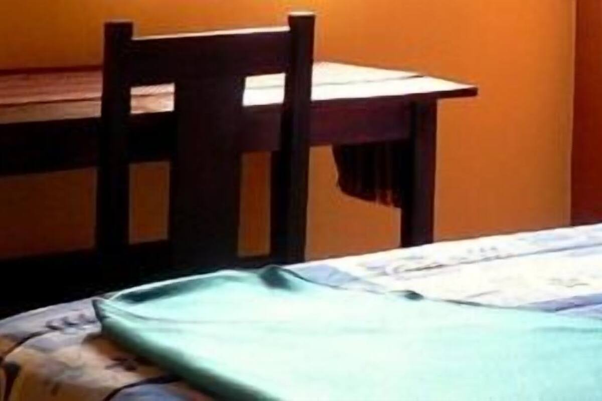 A bed with a patterned bedspread, a wooden chair, and a table against a wall.