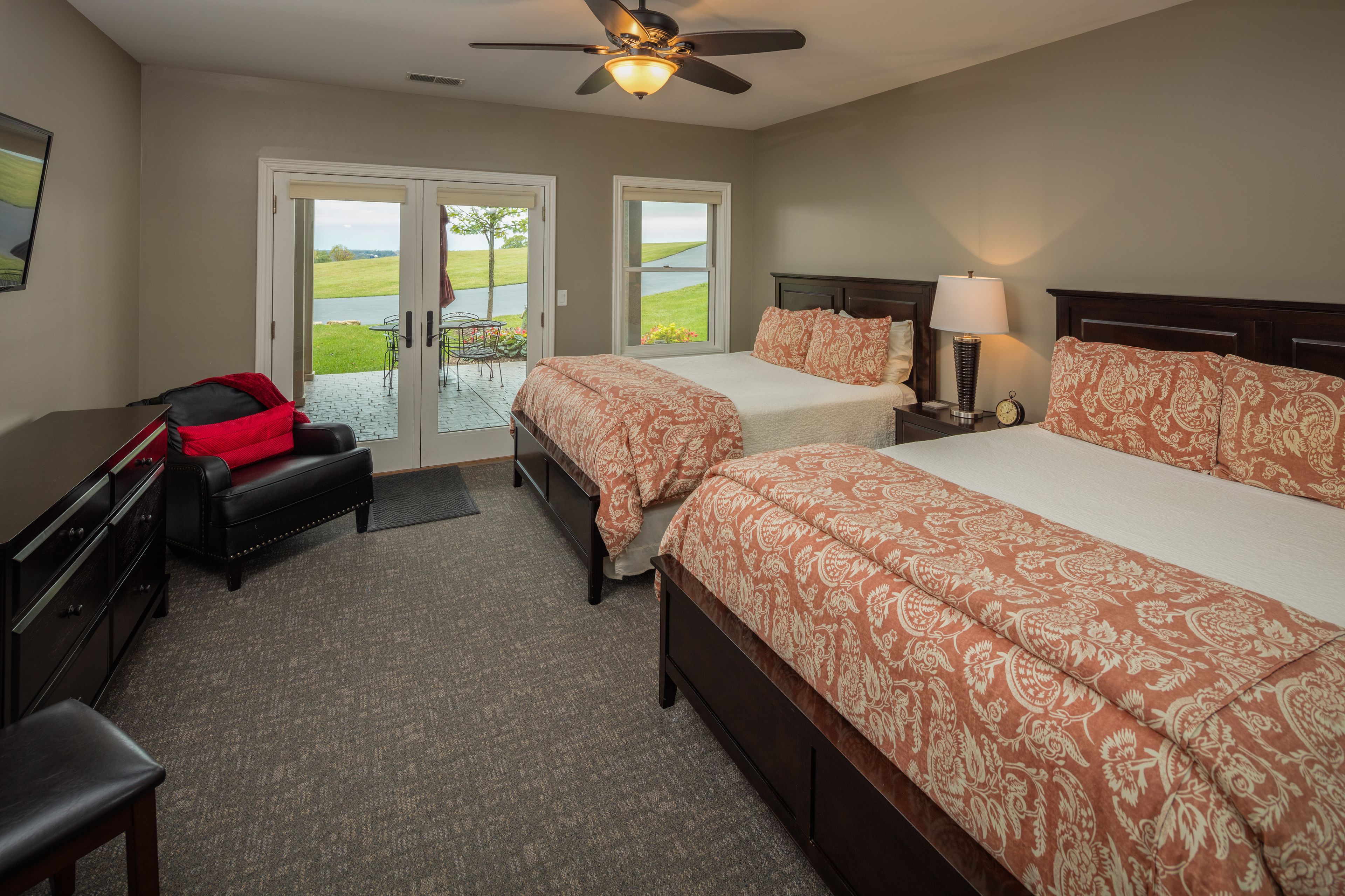 A bedroom with two beds, a desk, and a chair. There is a ceiling fan and a window with a view of a green landscape.