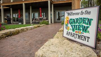 Une pancarte accueillante indique l’entrée de l’appartement « Garden View », avec un chemin en briques menant à un espace extérieur couvert aménagé avec du mobilier.