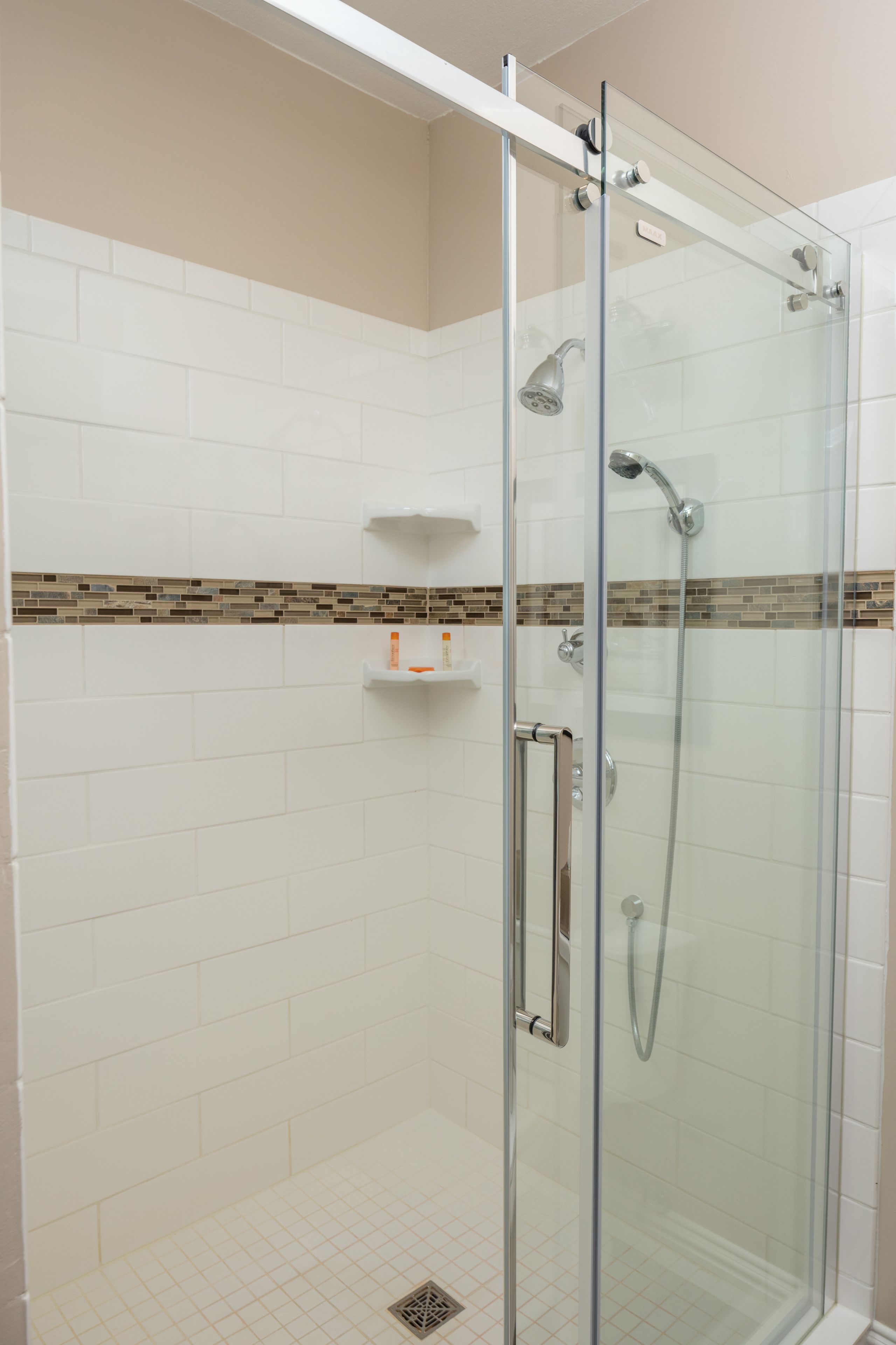 A glass-enclosed shower with a white tiled wall, a beige upper section, and a brown-tiled lower section.