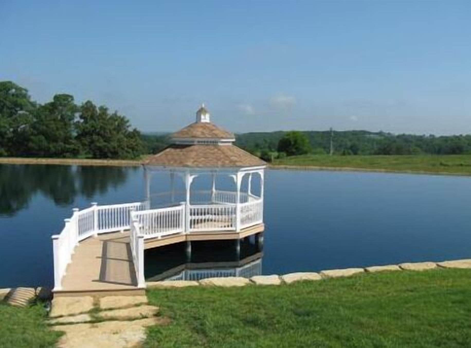 A gazebo with a white railing on a dock extending into a calm lake, surrounded by greenery.