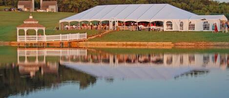 A large white tent with a covered walkway, set on a grassy area near a body of water.