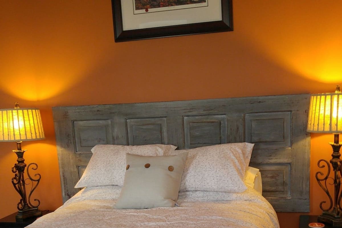 Ein ordentlich bezogenes Bett mit einem gemusterten Überwurf, zwei Nachttischlampen und einem gerahmten Bild an der Wand.