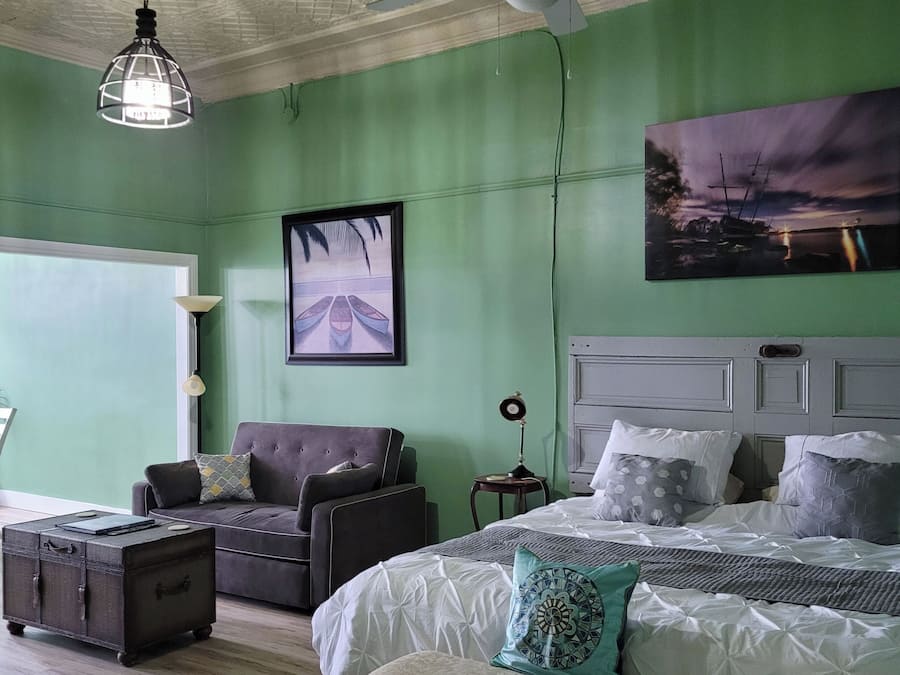 Un dormitorio con cama, un banco, una silla, una mesita y un ventilador de techo.