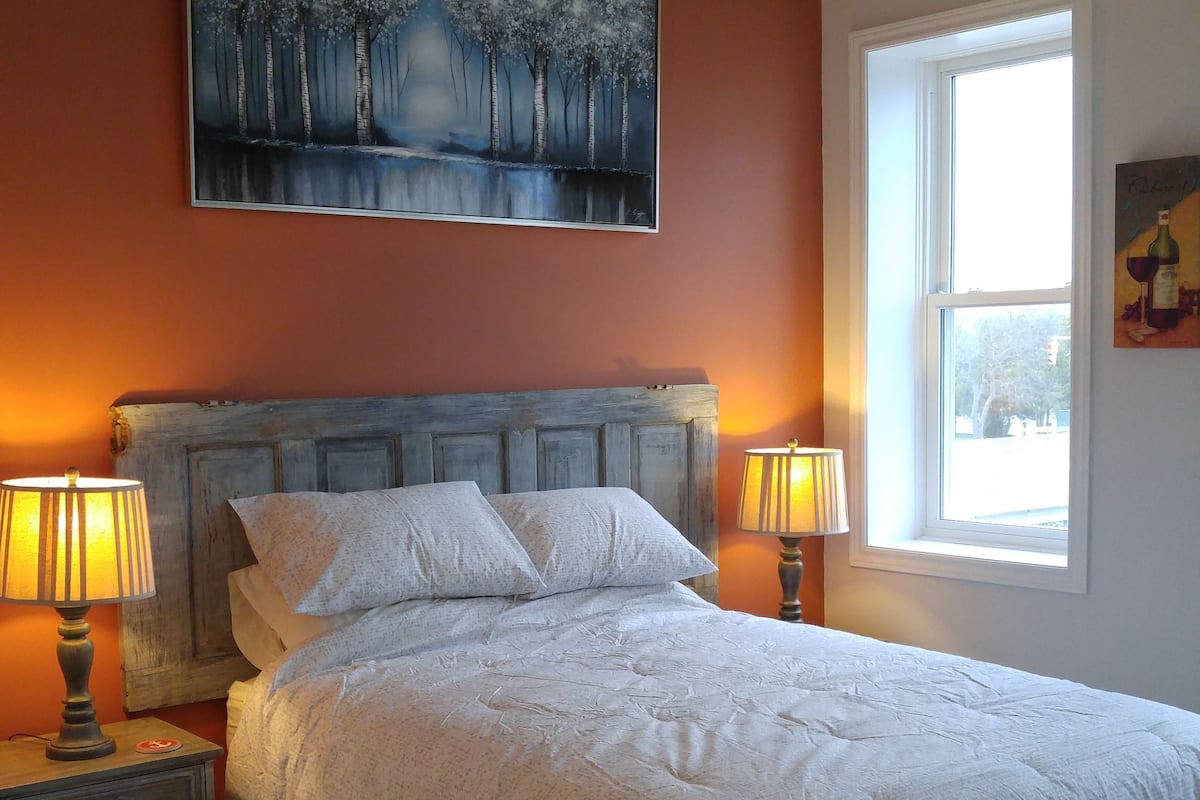 Ein Schlafzimmer mit einem großen Bett, zwei Nachttischlampen, einem Nachttisch, einem Sessel, einem Fenster und einem Bild an der Wand.