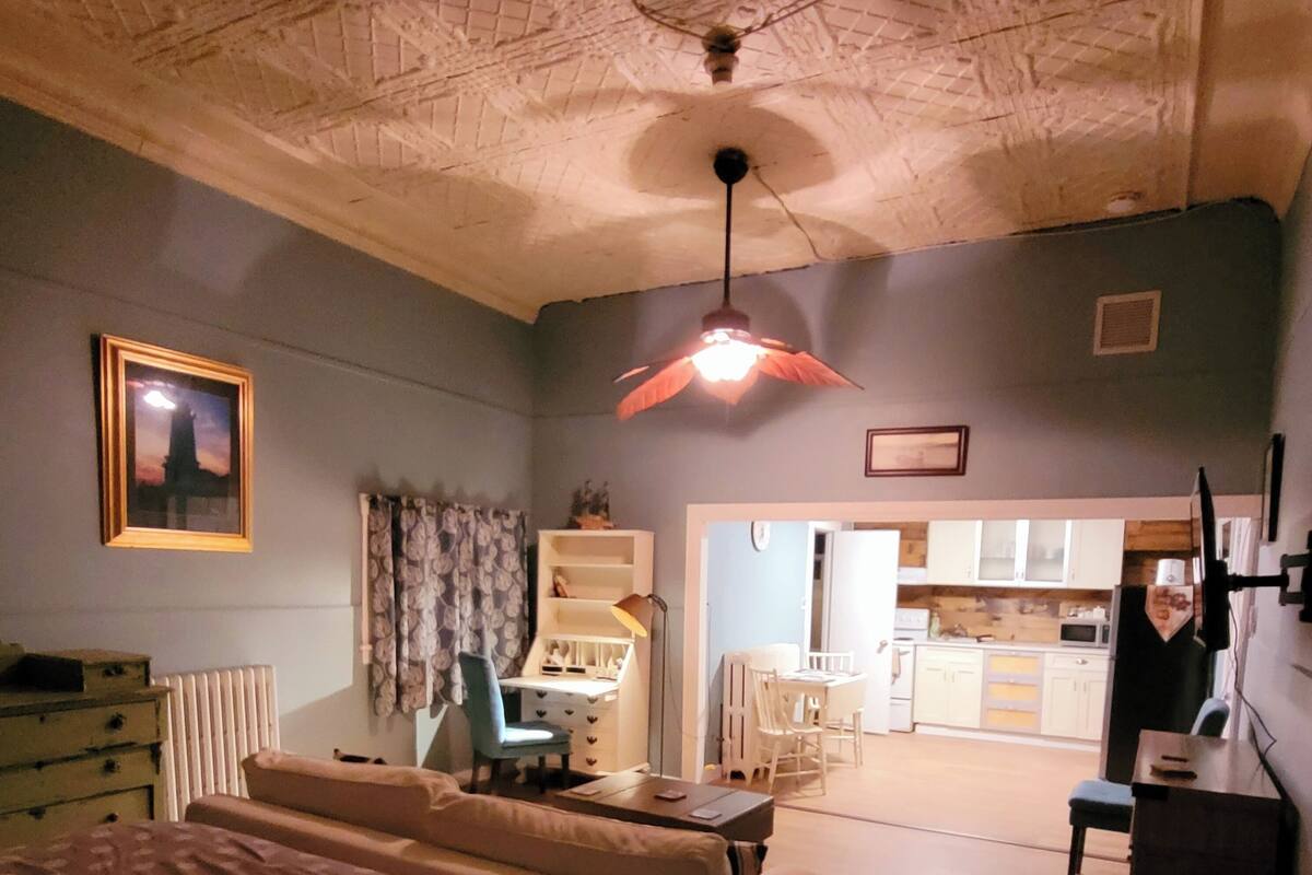 Ein Schlafzimmer mit einem Bett, einem Schreibtisch, einem Stuhl, einem Heizkörper und einer kleinen Küche im Hintergrund.