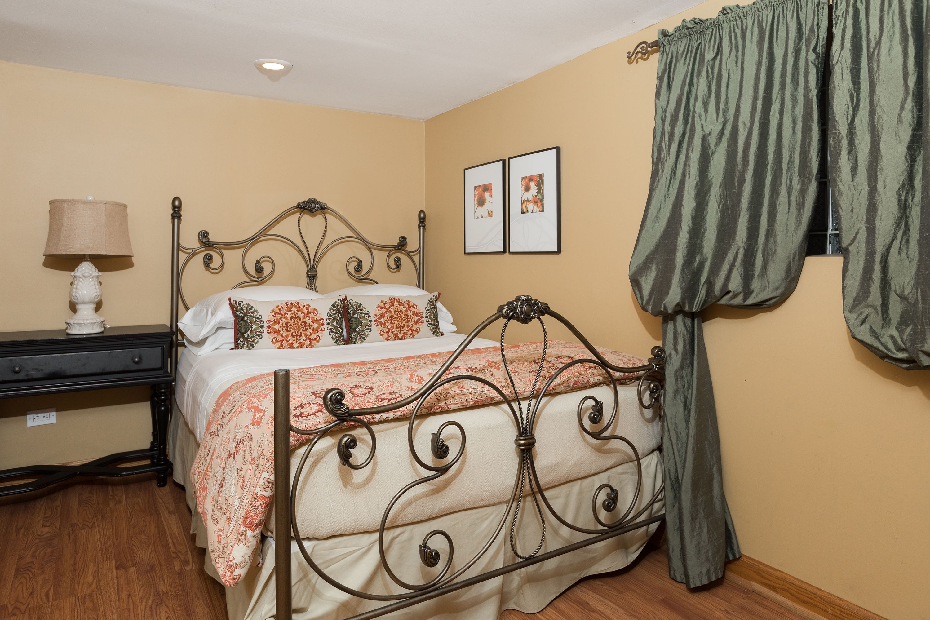 A bedroom with a metal bed frame, a bedside table with a lamp, and two framed pictures on the wall.