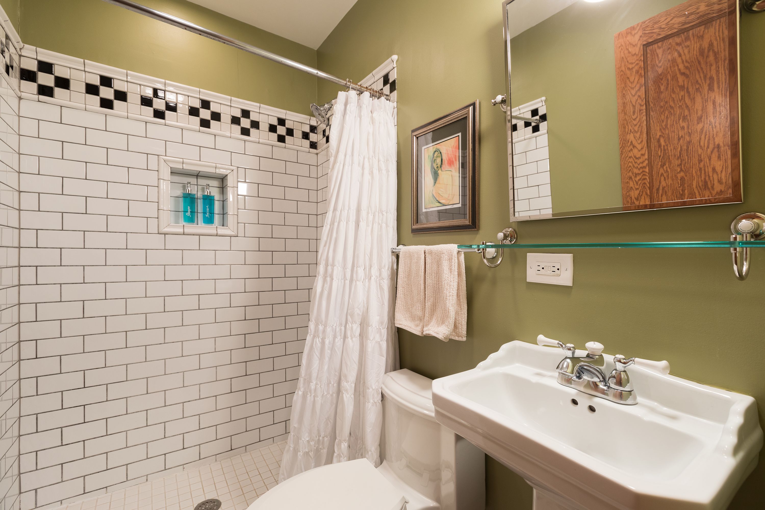 Un baño con paredes de azulejos blancos, un inodoro blanco, un estante de vidrio con artículos de aseo y un mueble de madera.