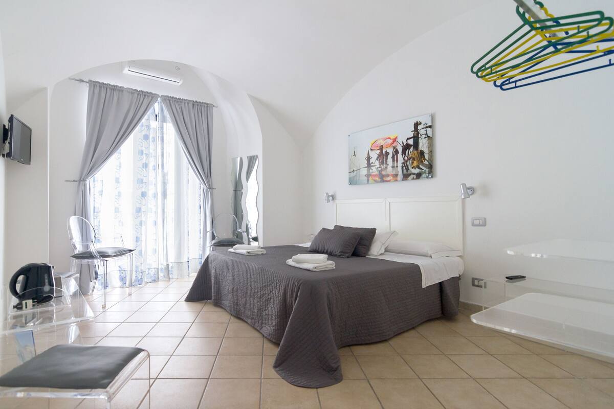 A modern hotel room with a bed, a chair, a desk with a kettle, and a window with curtains.