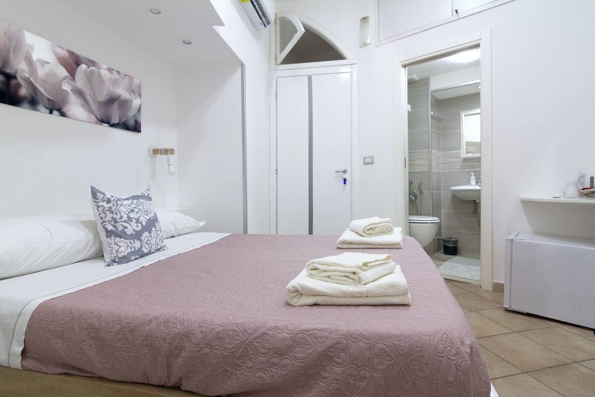 A bedroom with a bed, a bedside table, a nightstand, a door, and a bathroom with a shower and toilet.