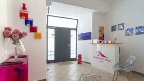 Un espace d’accueil avec un bureau, une chaise et un vase de fleurs. Des tableaux encadrés ornent le mur, et une porte donne sur l’extérieur.