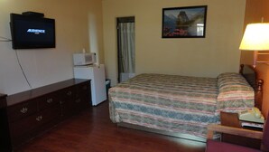 A hotel room with a bed, a television displaying 'AUDIENCE MUSIC', a microwave, a small refrigerator, a wooden dresser, a bedside table with a lamp, and a painting on the wall. - Sunset Motel (Taft)