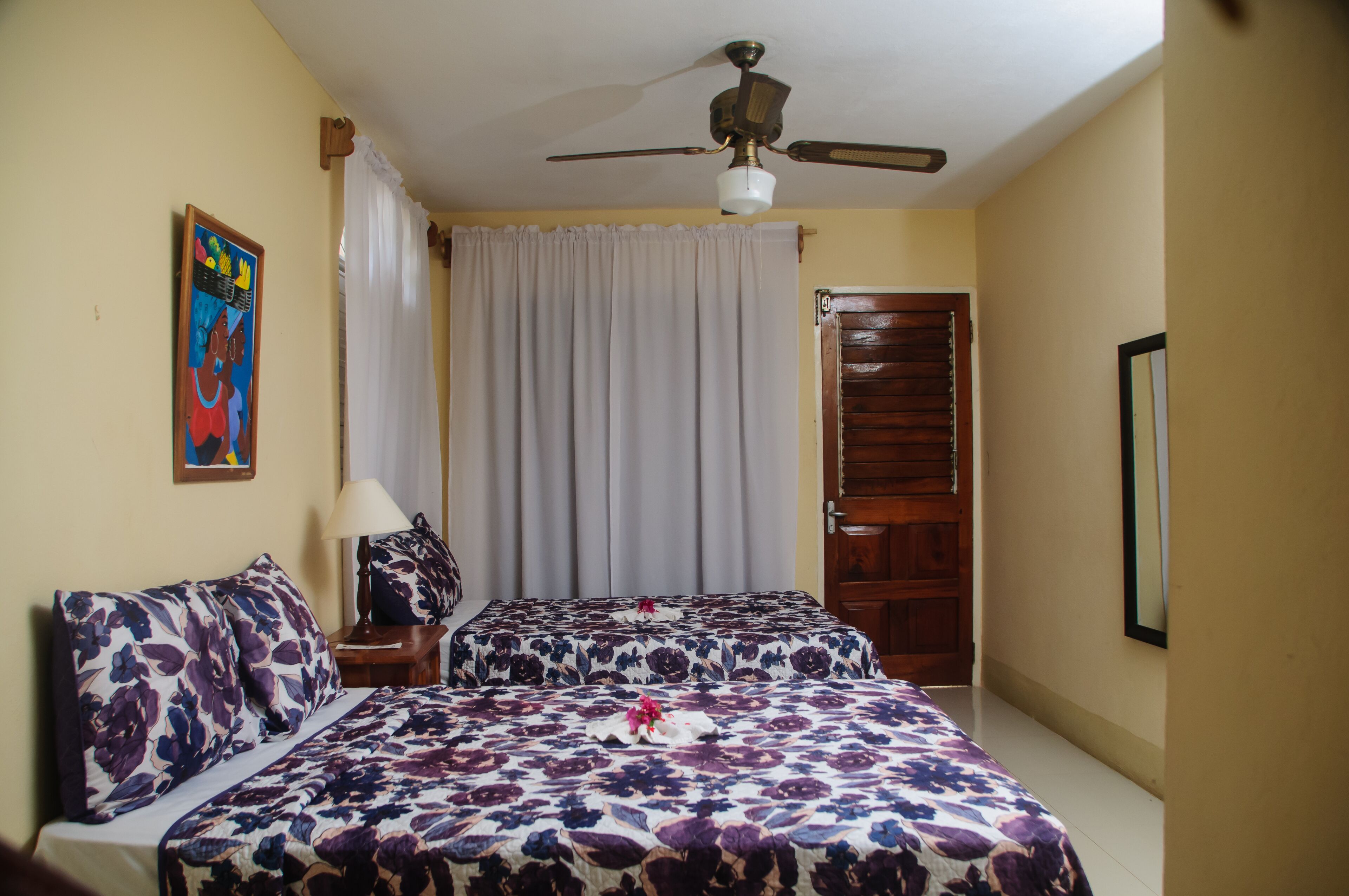 Une chambre avec deux lits, un ventilateur de plafond, une porte et un tableau au mur.