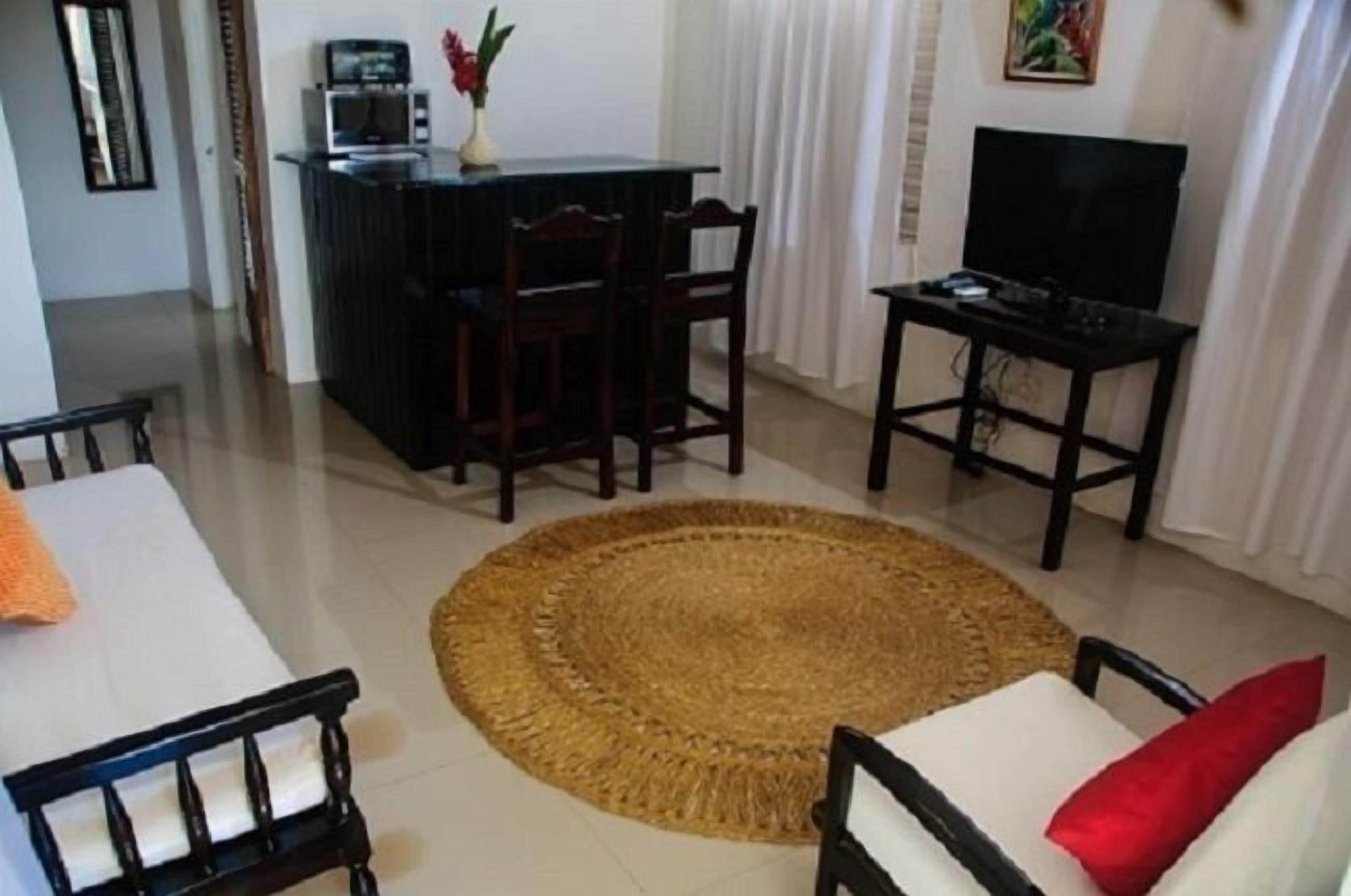 A living room with a sofa, a coffee table, a TV, a small kitchen area with a microwave, and a dining table with chairs.