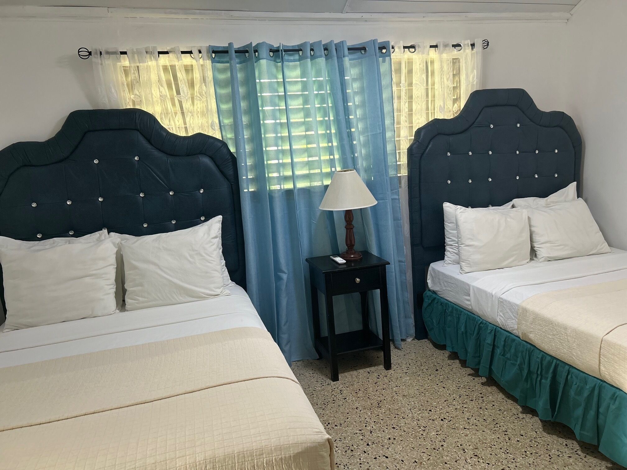 A hotel room with two beds, a nightstand with a lamp, and a window with blue curtains.