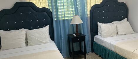 A hotel room with two beds, a nightstand with a lamp, and a window with blue curtains.