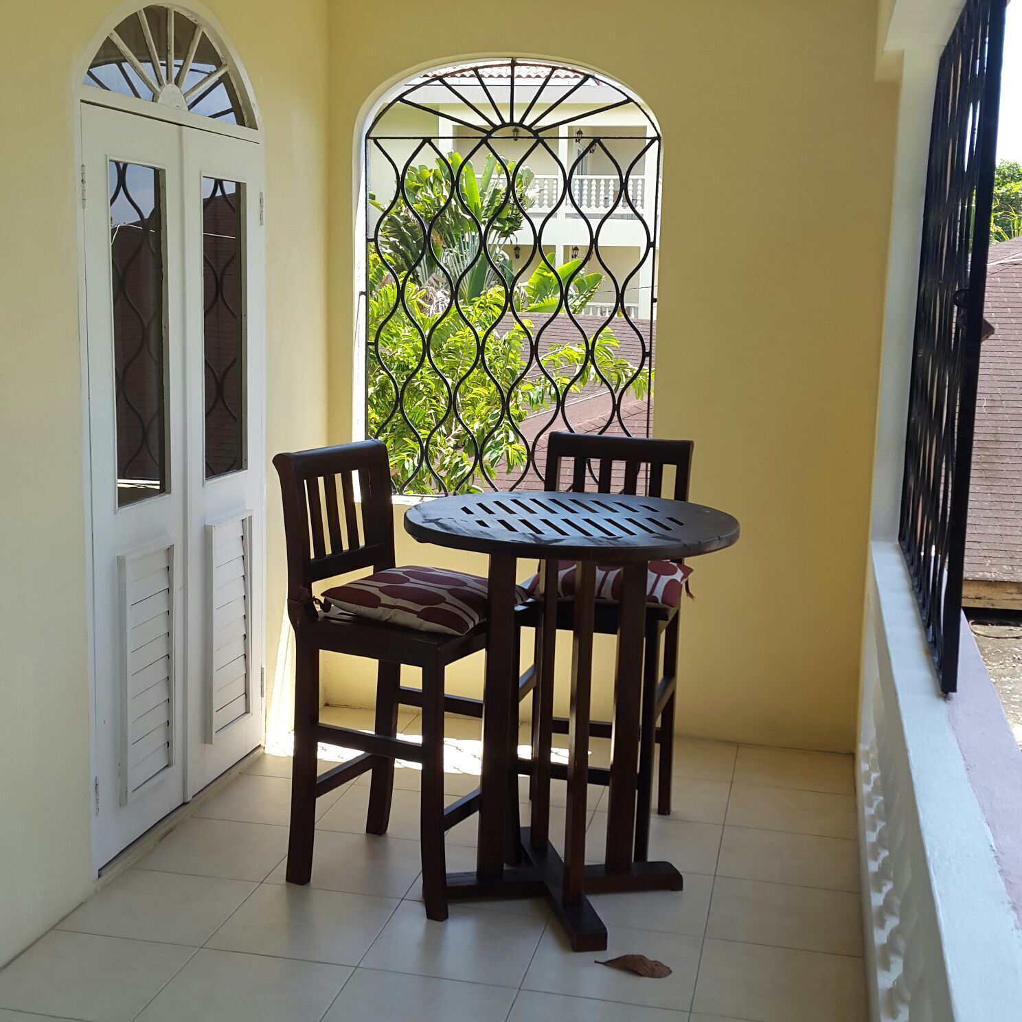 A small balcony with a round table and two chairs, a white door with glass panels, and a metal gate with a view of greenery.