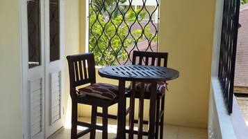 A small balcony with a round table and two chairs, a white door with glass panels, and a metal gate with a view of greenery.