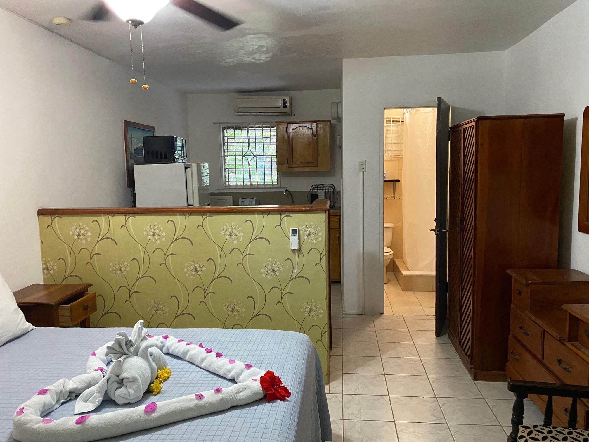 Quarto de hotel com uma cama, cômoda de madeira, uma mesinha e uma cozinha compacta ao fundo.