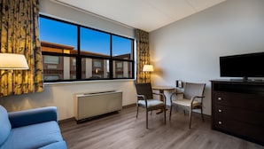 A hotel room with a large window, a blue sofa, two wicker chairs, a small table, a TV, and a dresser. - Gateway Inn (Merrick)