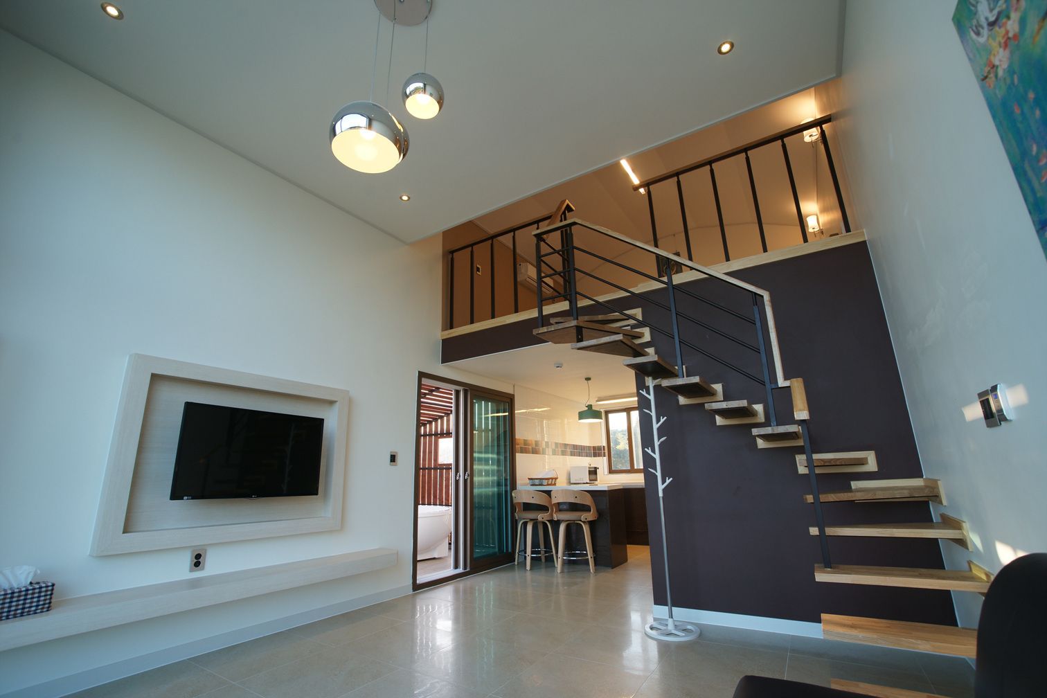 A modern interior with a staircase, a flat-screen TV mounted on the wall, and a kitchen area visible through an open door.