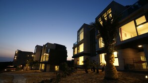 A modern building with multiple floors and large windows, surrounded by a sandy landscape with palm trees. - Donnaekohill Resort (Seogwipo)